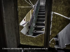 Durch eine zerbrochene Glasscheibe sieht man eine alte Treppe in diesem verlassenen Gebäude.