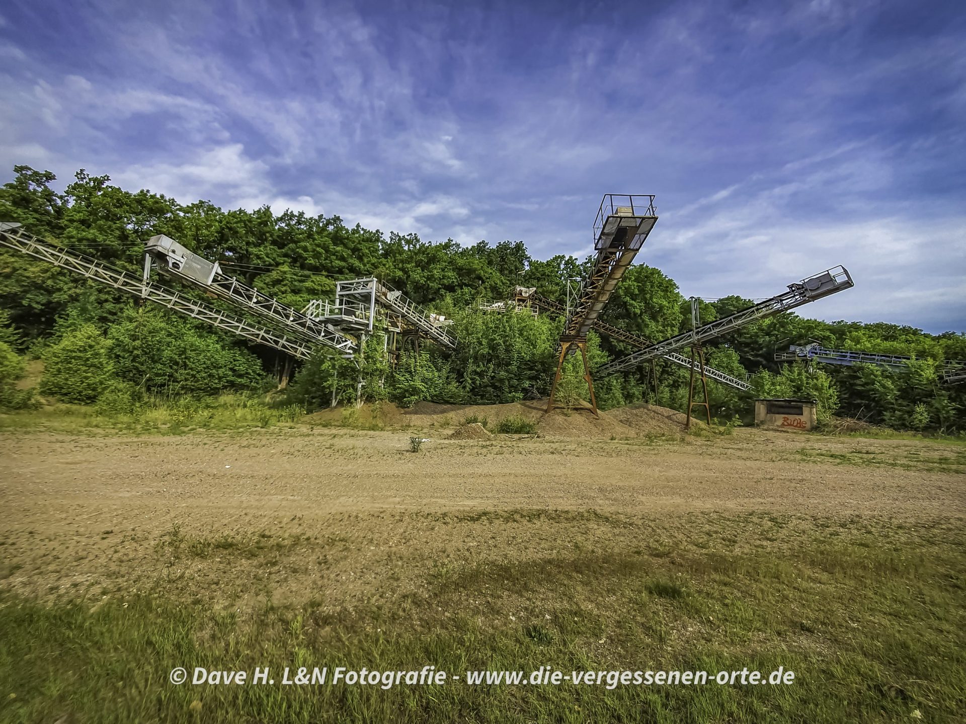 Auf dem Platz die großen Fördermaschinen von dem verlassenen Kieswerk, im Hintergrund die Bäume de umgebenden Waldes.