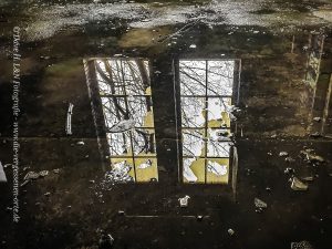 Eine Wasserspiegelung, ein Fenster erstrahlt hell in der Pfütze einer verlassenen Industrieanlage.