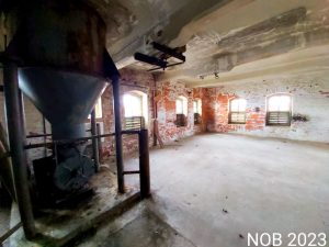Eine weitläufige Lagerhalle der Ölmühle, vorne links ein verrostetes Fallrohr das von der Decke ragt.
