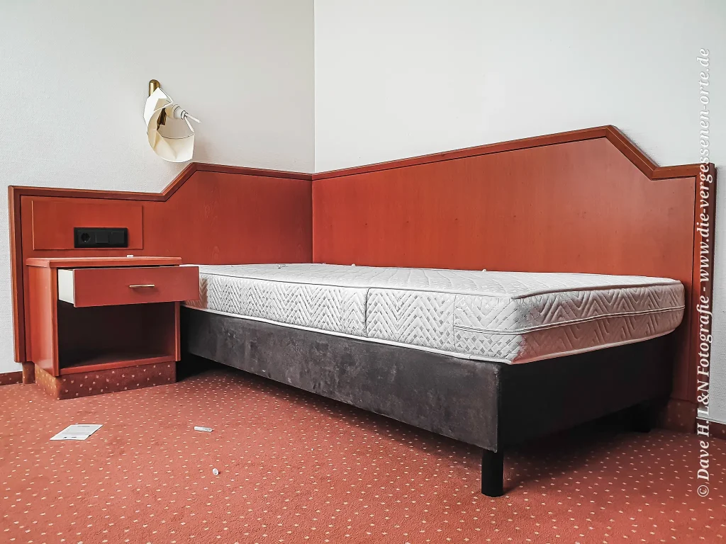 Bett in einem verlassenen Hotelzimmer, darüber eine zerschlagene Lampe.