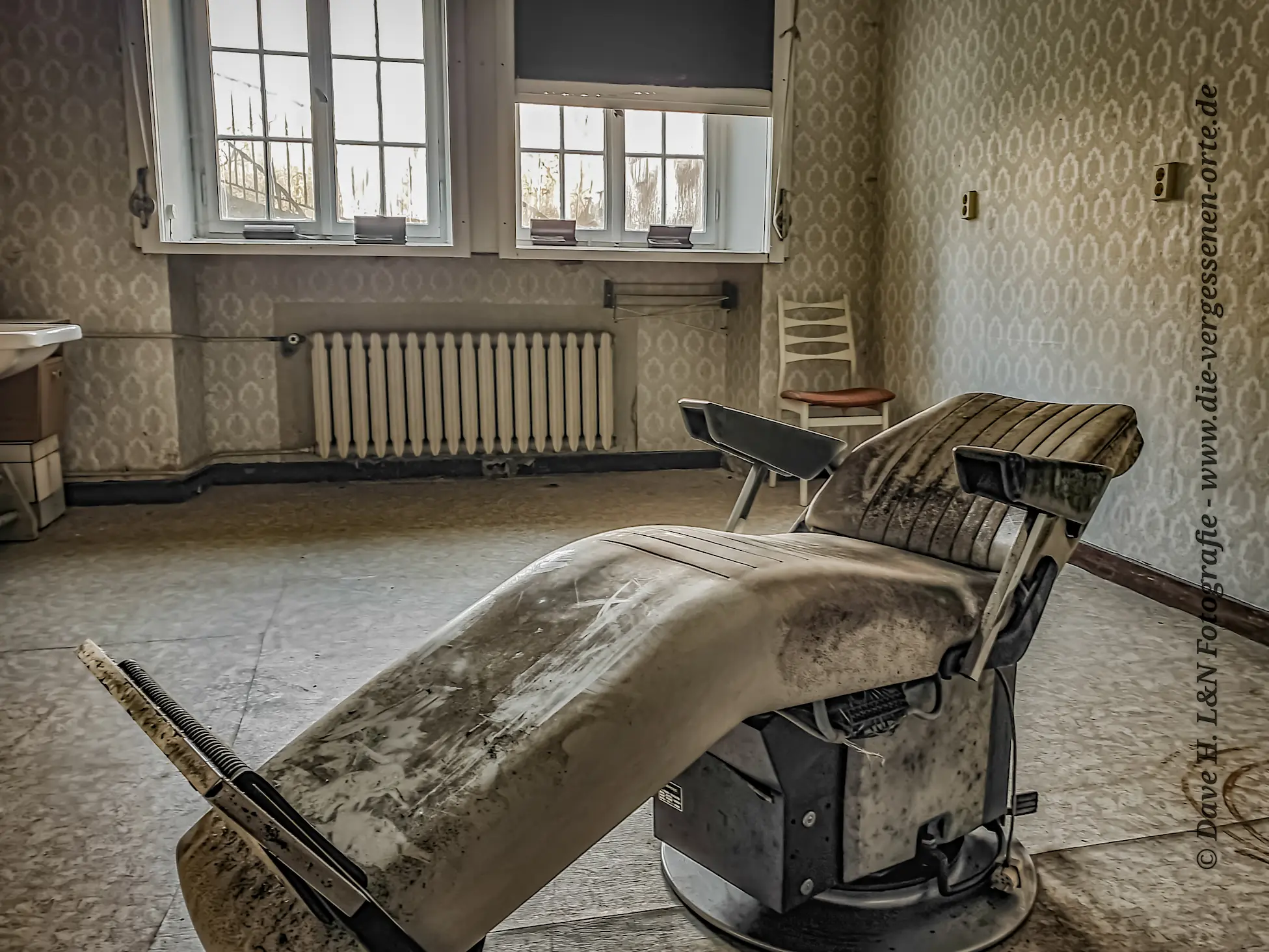 Ein alter, verlassener Behandlungsstuhl in einem verfallenen Raum mit Fenster und einer alten Tapete.