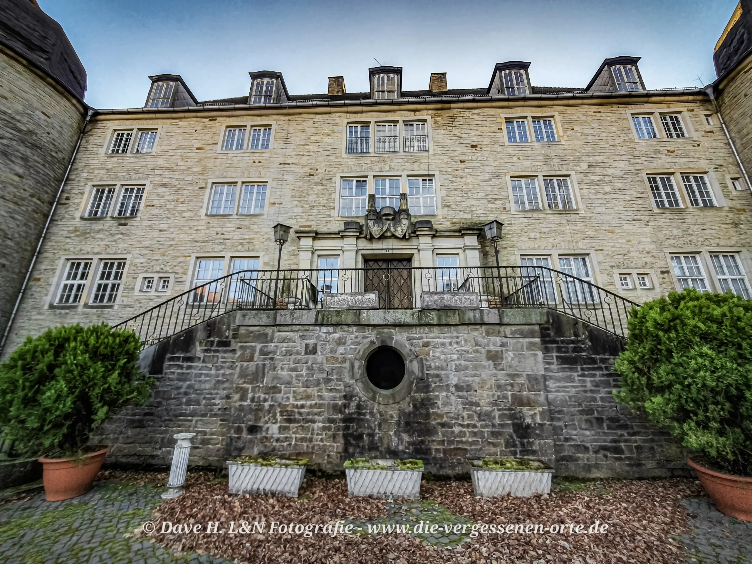 Fassade eines verlassenen Schlosses mit Treppenaufgang und großen Fenstern, umgeben von Bäumen und Pflanzkübeln.